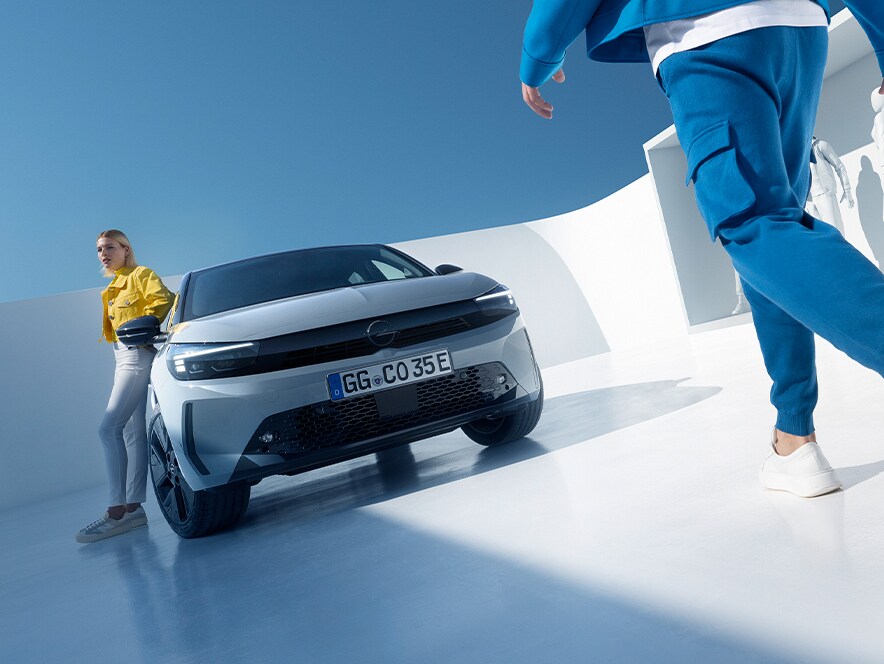 Front view of a grey Opel Corsa Electric with black alloys, woman leaning against the door and man walking towards them