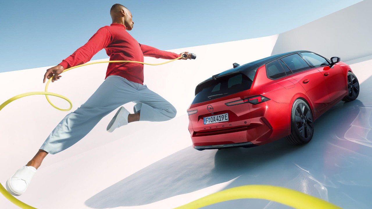Ein Mann springt mit einem EV-Ladekabel in die Luft, im Hintergrund ein Opel Astra Sports Tourer Electric