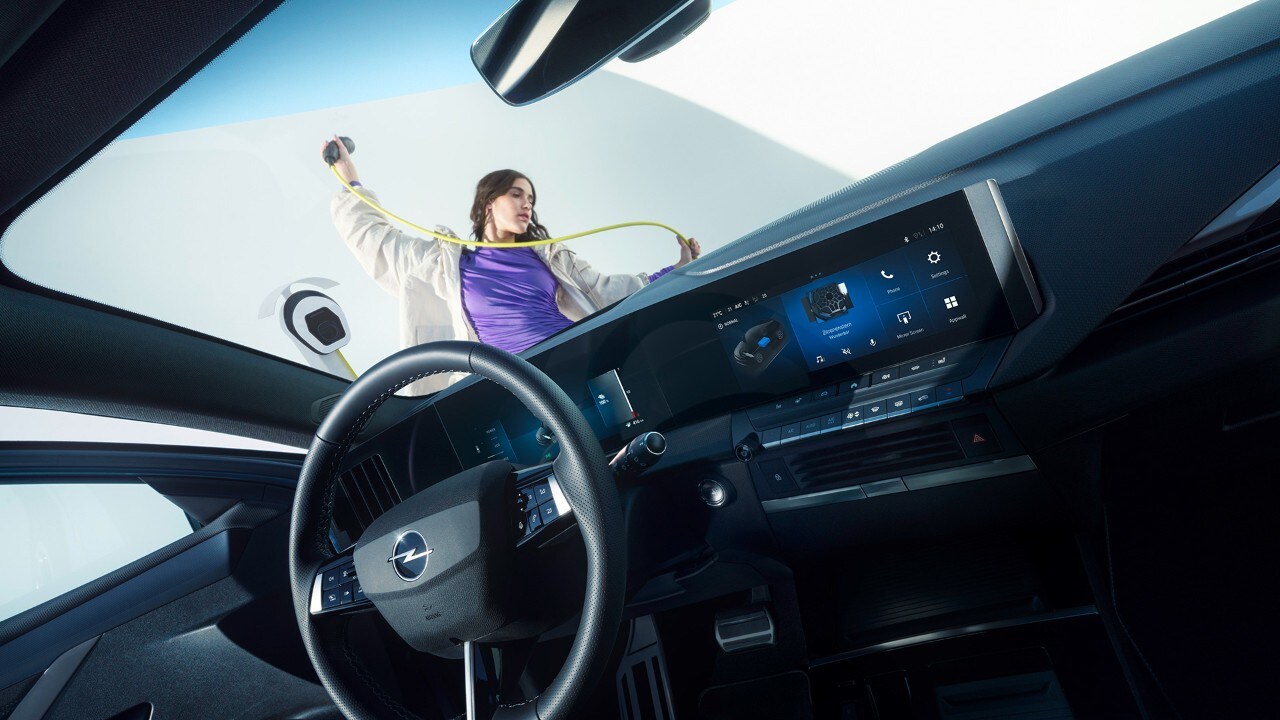 Innenansicht eines Opel Astra Electric vom Beifahrersitz aus und einer Frau, die draußen ein Ladekabel hält